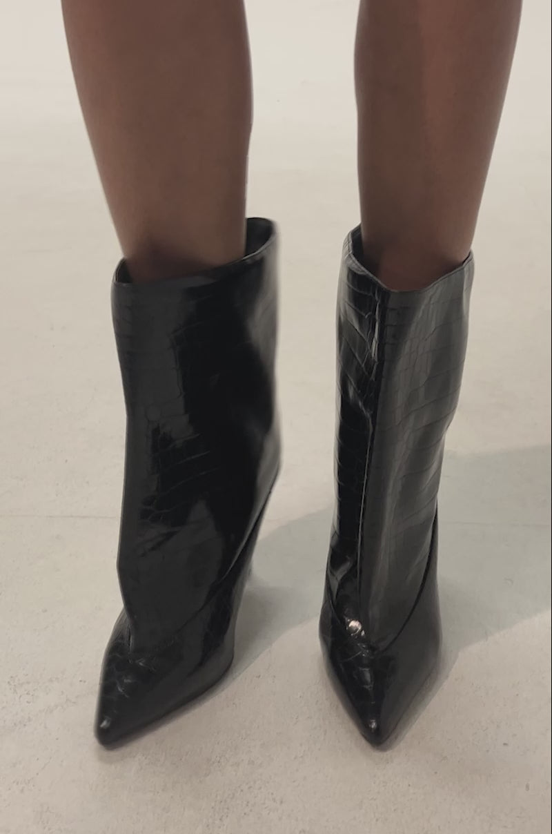 Video of woman modeling the AKIRA GOLD RITUAL BLACK CROC STILETTO BOOTIE—shiny black, crocodile-textured, pointed-toe stilettos with a wide shaft that reaches mid-calf—styled with bare legs against a plain background.