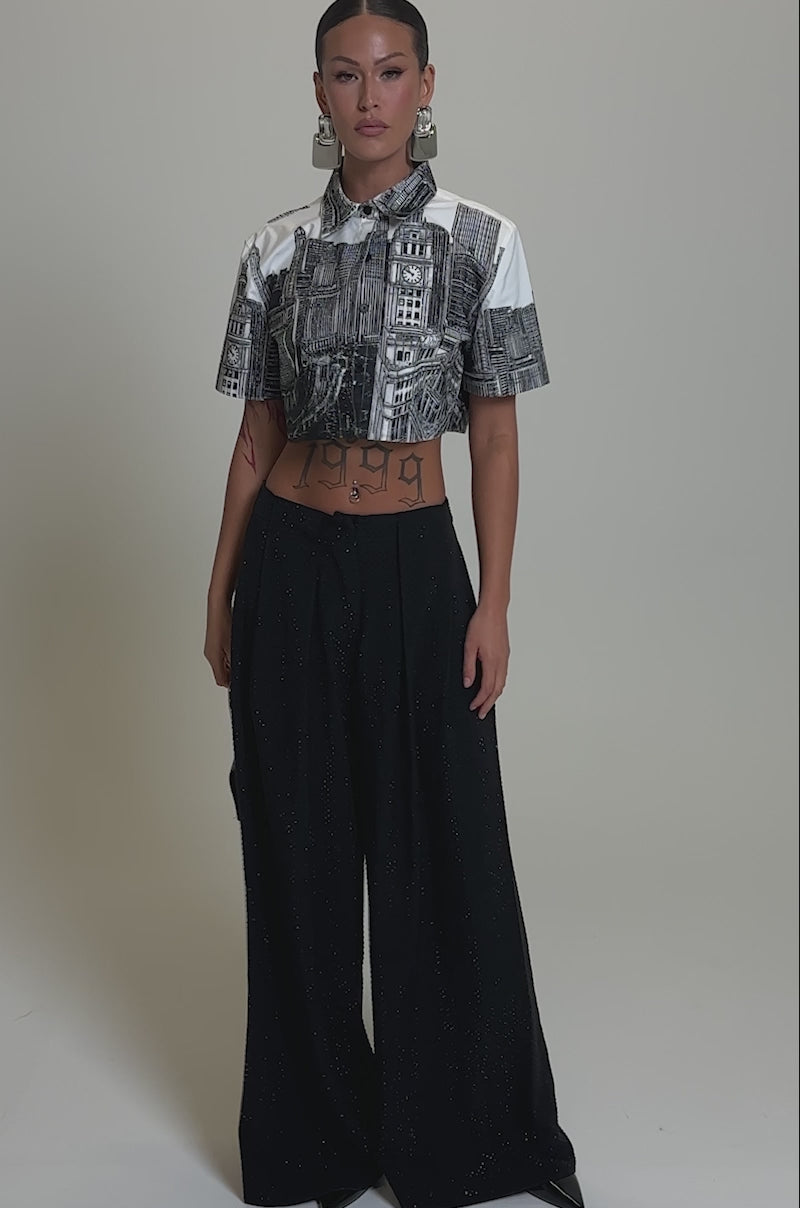 A video of a model wearing the SPARKLE QUEEN TROUSER, with wide-leg black pants and a cropped cityscape-print shirt,  with statement earrings, a small handbag, and sleek hair.