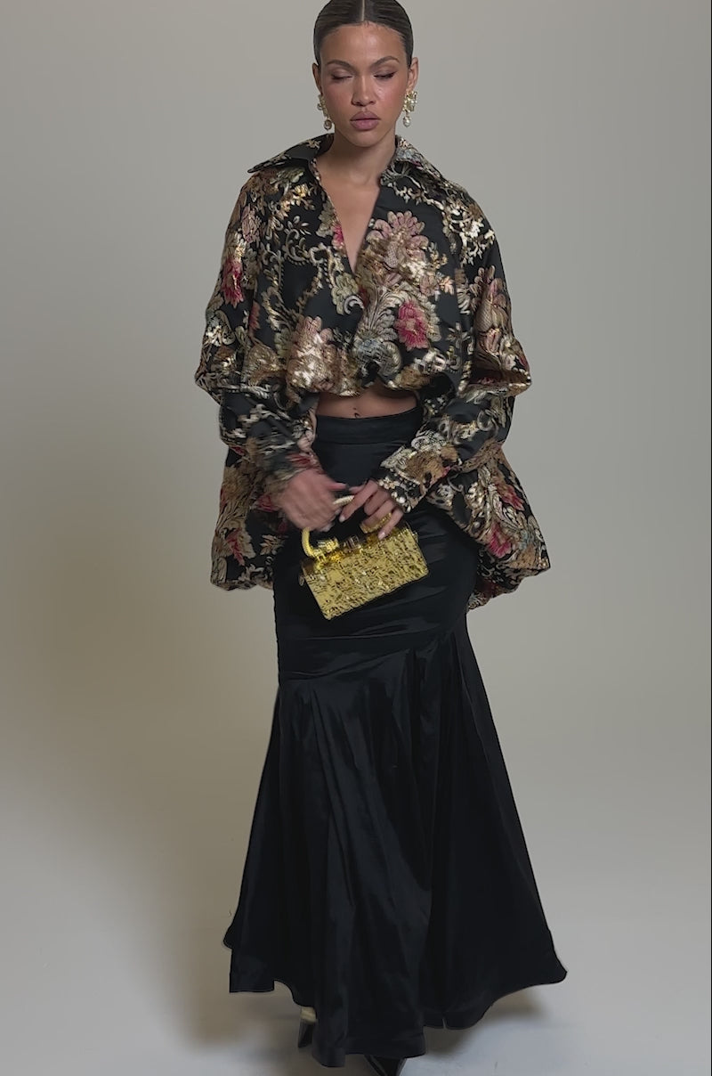 Video of woman modeling the HIGH SOCIETY HIGH LOW BROCADE BLOUSE—a gold-tone metallic top with floral patterns—paired with a black satin skirt, gold earrings, and a small gold bag, her hair tied back.