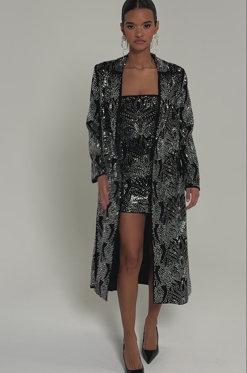 Video of woman modeling the SHE IS THE MOMENT RHINESTONE TRENCH with shimmering silver patterns. She has sleek pulled-back hair, large earrings, and black pointed-toe heels.
