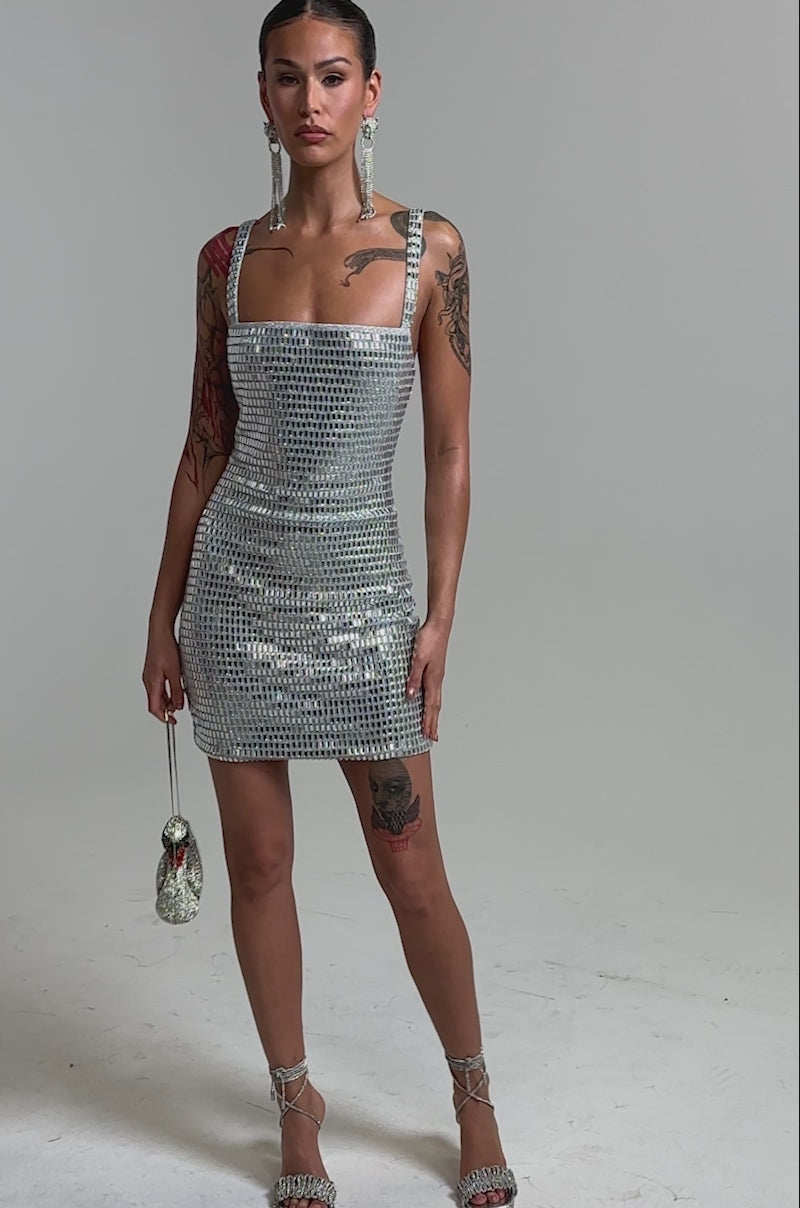 Video of woman modeling the BAILEY RHINESTONE MINI DRESS, a sleeveless silver sequin party dress, holding a swan-shaped handbag. She accessorizes with long earrings and shows visible tattoos, standing against a plain neutral background.