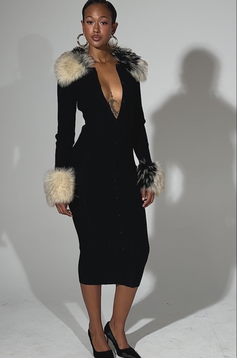 A video of a woman standing confidently in the MICHELLE KNIT MIDI DRESS WITH FUR ACCENTS, featuring a ribbed knit design, deep V-neckline, front buttons, and faux fur on the cuffs and collar. She pairs it with hoop earrings and black heels against a light background.