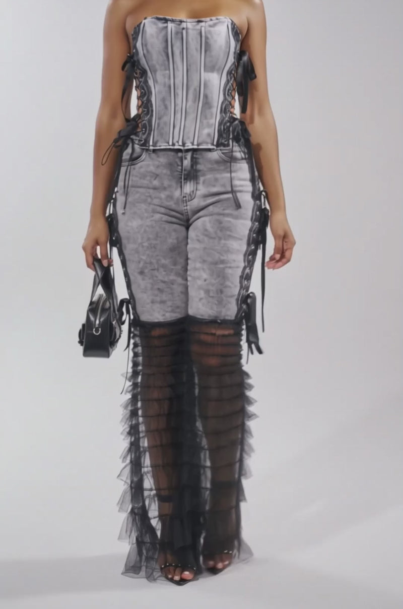A video of a woman in the TICKED AND TIED LACE DETAIL FLARE DENIM—high-rise denim pants with sheer black ruffled panels and black lace-up sides—paired with a gray corset top, black heels, and a small black handbag.