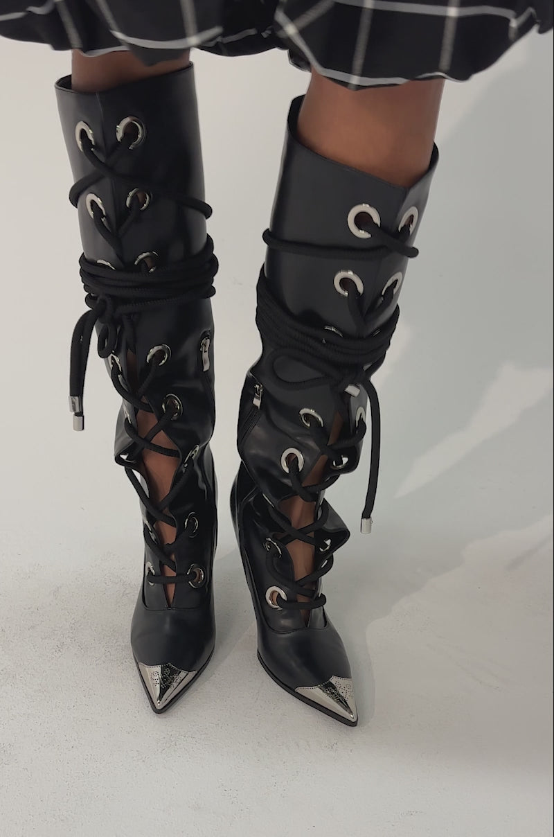 Video of woman modeling the AKIRA GOLD PANTOUM BLACK PU HARDWARE BOOT—black faux leather, knee-high stilettos with metallic pointed toes