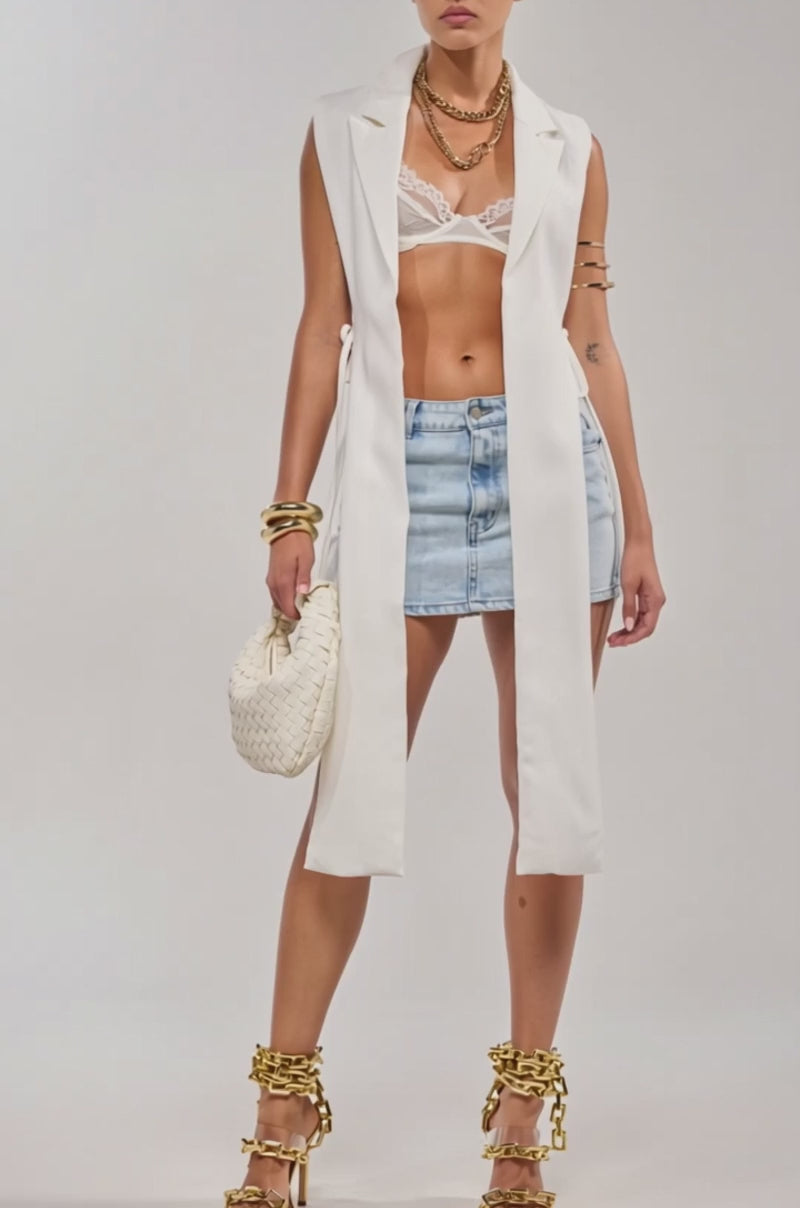 A video of a woman in the HONEY HONEY TIE VEST with a notch lapel collar, white lace bra, light blue denim mini skirt, gold jewelry, chain-detailed gold-heeled sandals, and a woven white handbag against a plain background.