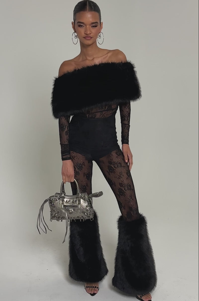 Video of woman modeling the DELARA FUR TRIM LACE JUMPSUIT, featuring sheer black lace pants and matching faux fur leg warmers. She accessorizes with a metallic handbag and large hoop earrings, showcasing bold elegance.