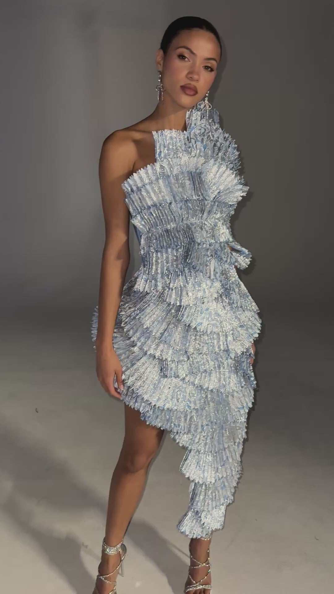 A video of a model wearing the LUST FOR LIFE ASYMMETRICAL BROCADE MIDI DRESS IN BLUE.  Featuring a stretchy bandage base, a one-shoulder neckline, pleated brocade ruffle paneling with stunning metallic detailing lining the front, and a discreet side zip closure. Complete with an asymmetrical hem for a sculptural, refined finish that turns every entrance into a moment. 