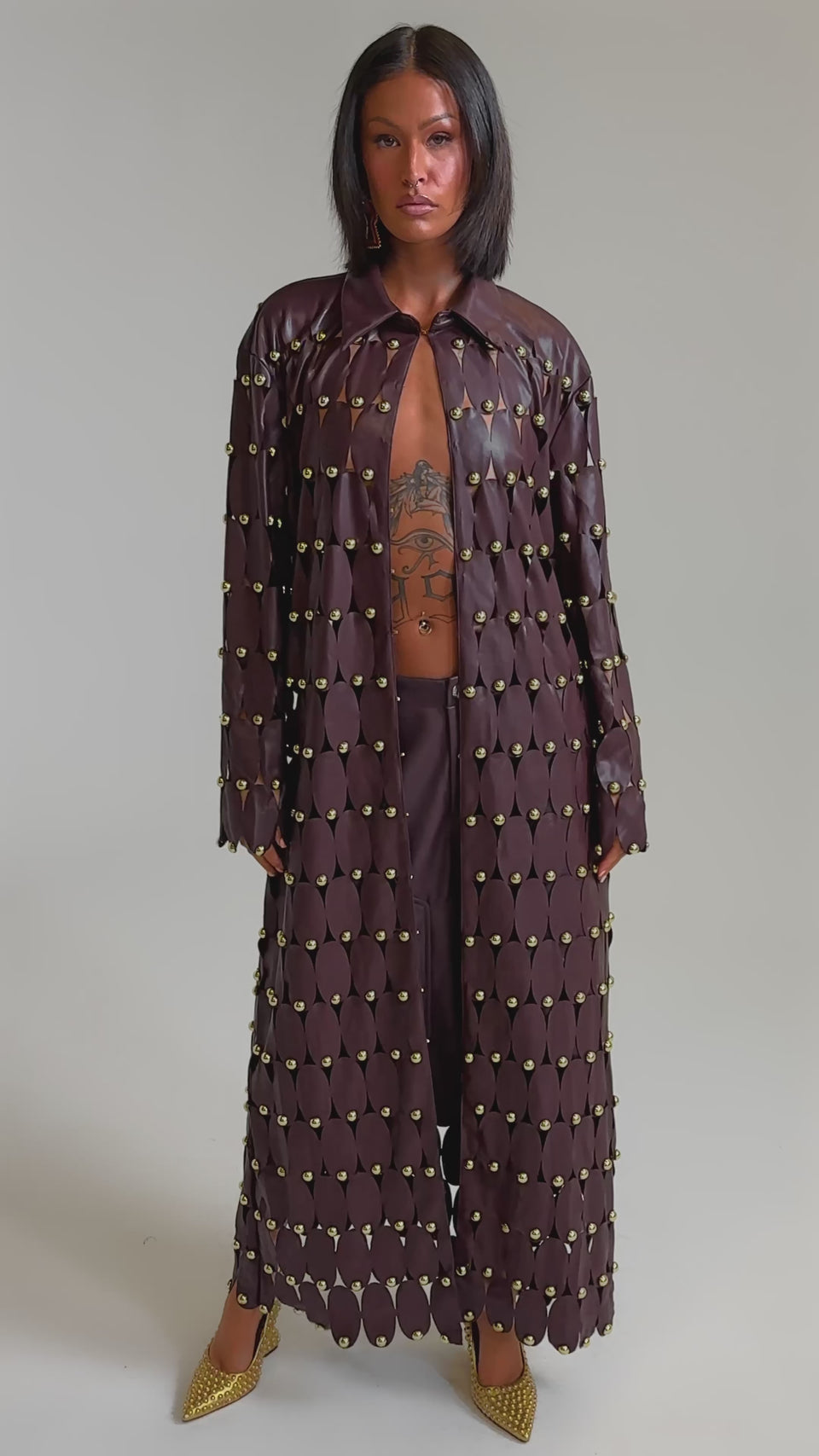 A video of a woman wearing THE SNEAK PEEK STUDDED PU LONG CARDIGAN IN BURGUNDY over a matching outfit, showing tattoos on her chest and abdomen and finishing the look with gold pointed heels.