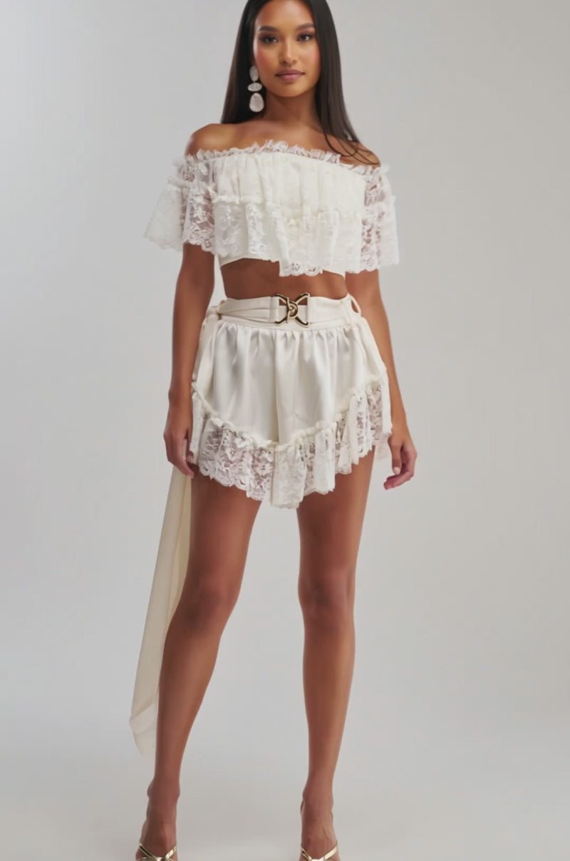 Video of woman modeling the PRIMROSE LACE BLOUSE and high-waisted cream shorts with a gold belt, standing against a plain background.