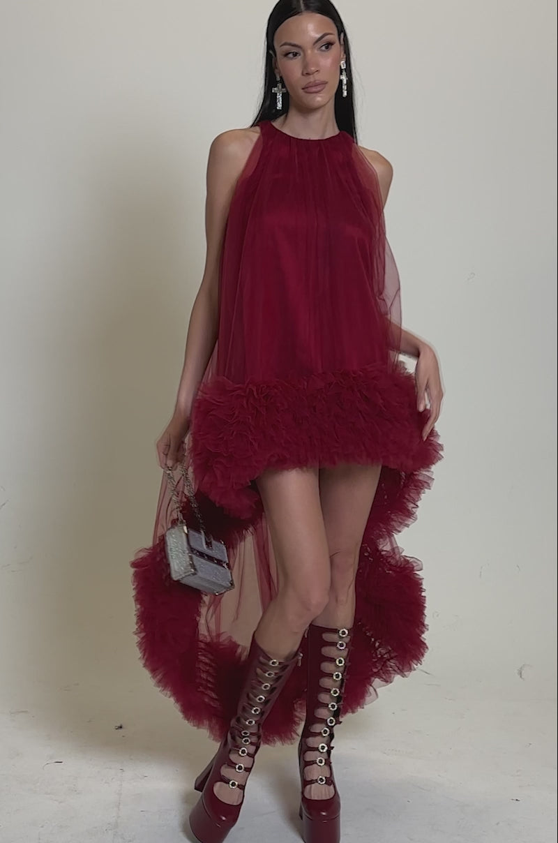 A video of a woman in the ARI TULLE HIGH LOW DRESS IN BURGUNDY by AKIRA Label, styled with tall lace-up red platform boots and a small handbag.