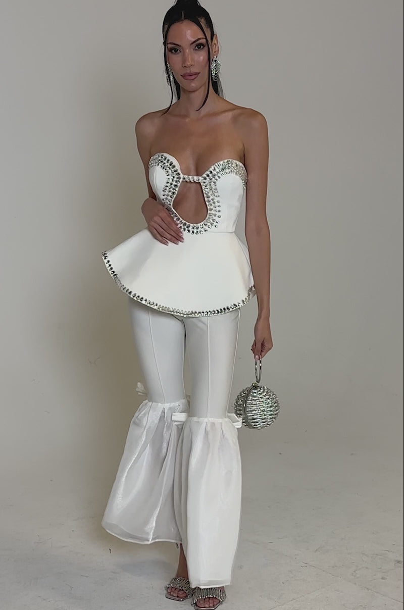 A video of a woman posing against a white background wearing the BLING BLING CORSET—a stylish white peplum corset top with front cut-out and silver embellishments—paired with fitted pants, sheer flared bottoms, silver heels, and a round silver handbag.