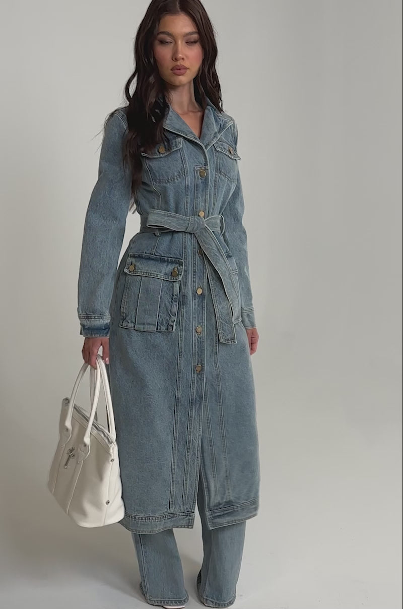 A video of a woman models the CAMILLA DENIM TRENCH, a long belted denim coat with cargo pockets, styled over matching denim pants and accessorized with a large white handbag background.