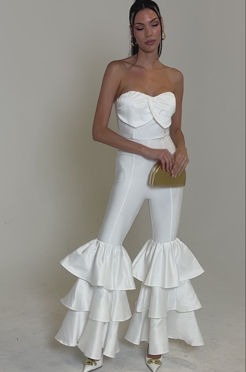 A video of a woman posing against a light background wearing the I JUST WANNA DANCE FLARE LEG JUMPSUIT IN WHITE. She holds a beige clutch, wears pointed-toe heels, and her straight hair is tucked behind her ears.