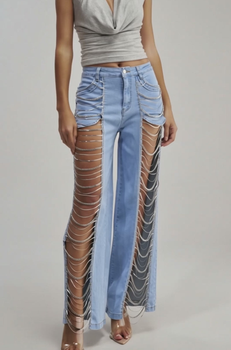 A video of a woman wearing the CHAINS FOR DAYS RELAXED DENIM—light blue cutout jeans with draped silver chains—and a sleeveless gray halter top, paired with clear high-heeled sandals.