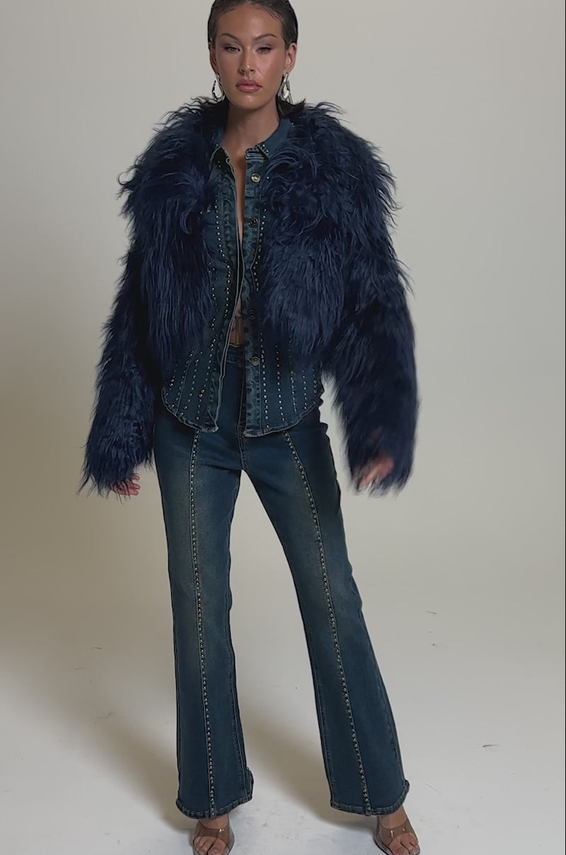 A video of a woman in the LIFE OF THE PARTY FAUX FUR jacket over a studded denim shirt and flared jeans with visible seams, paired with open-toe heels and hoop earrings against a plain background.