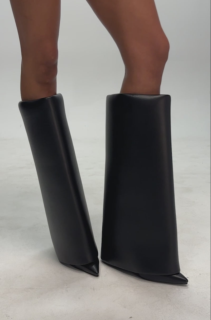 Video of a woman modeling the AZALEA WANG SALEM BLACK FOLD OVER WEDGE BOOT: black faux leather wedge boots with a pointed toe and wide, folded-over shafts reaching to the knees.