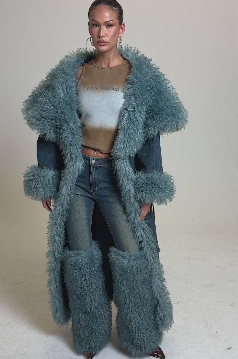 A video of a woman in the MOXIE FUR TRIM DENIM TRENCH, styled over distressed jeans and a cropped brown top. With slicked-back hair, hoop earrings, and a plain gray background, her look is both modern and bold.