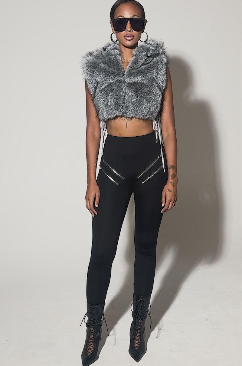 Video of woman modeling the TOUCH ME FUR TOP, a sleeveless gray cropped faux fur vest. She pairs it with large black sunglasses and zippered black pants against a light background.
