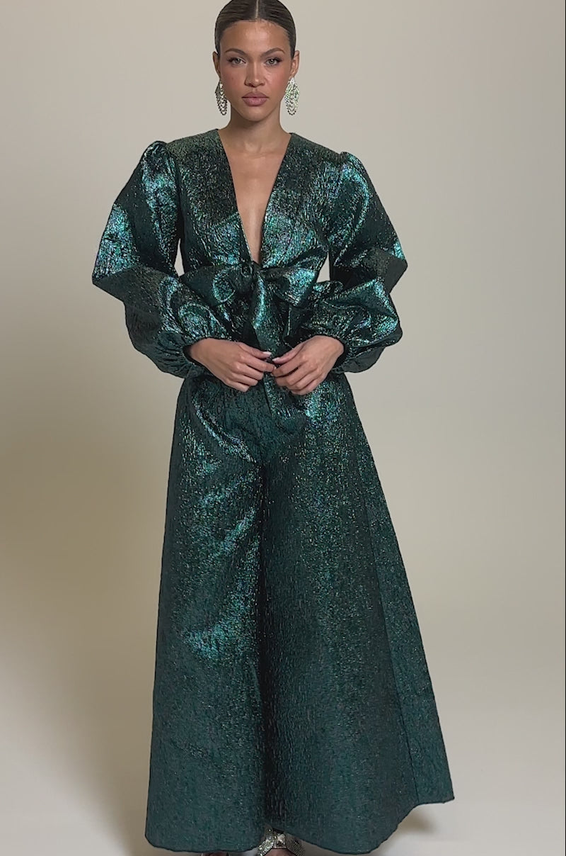 Video of woman modeling the ITS TIME TO CELEBRATE PALAZZO PANT, a teal brocade high-rise wide-leg trouser, styled with a deep V-neck top with a bow waist and puffed sleeves, plus heeled sandals and statement earrings.
