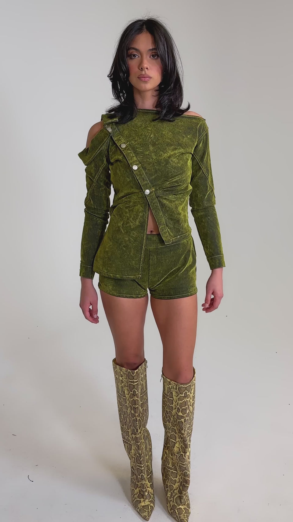 A video of a woman in a green AKIRA Label long-sleeve velvet top with cut-out shoulders, YOUR TIME IS COMING VELVET DENIM SHORTS, and knee-high snakeskin boots against a plain light background.