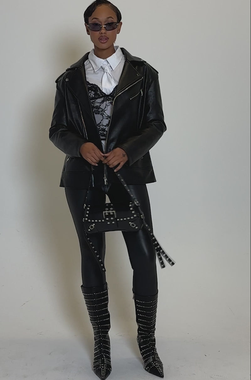 Video of woman modeling the THE BIGGER THE BETTER OVERSIZED MOTO women's faux leather jacket over a white shirt and black lace corset, paired with sunglasses and a black belt