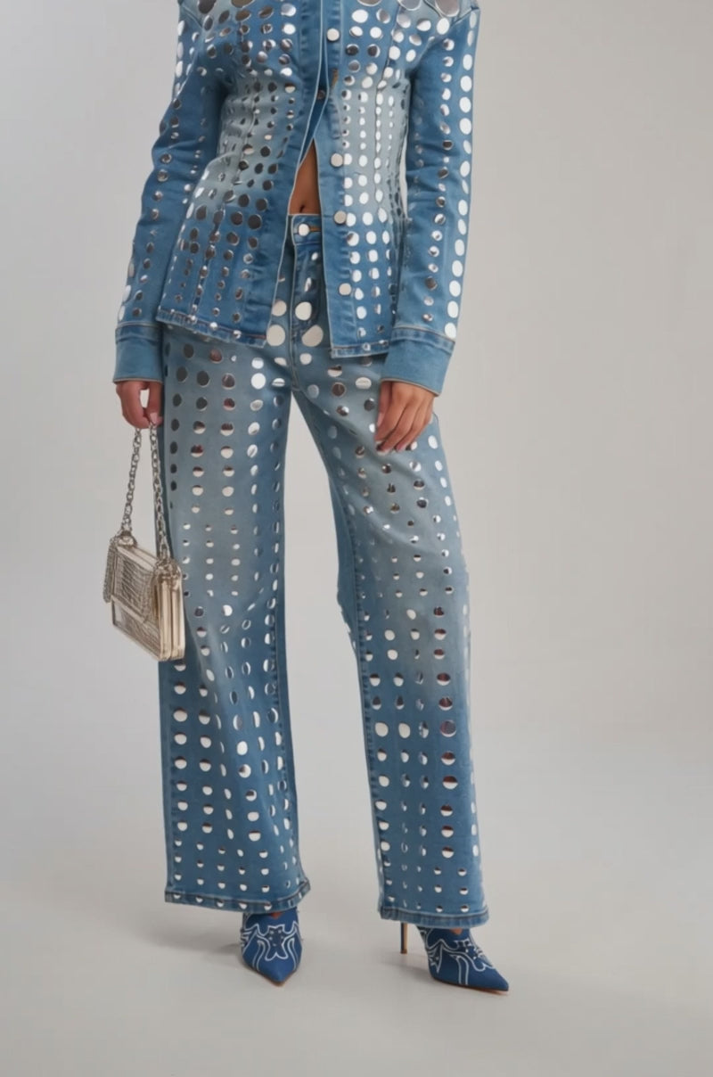Video of woman modeling the STUDS EVERYWHERE BUTTON DOWN denim top and matching pants set with large circular cutouts, a small transparent handbag, and blue high-heeled boots.
