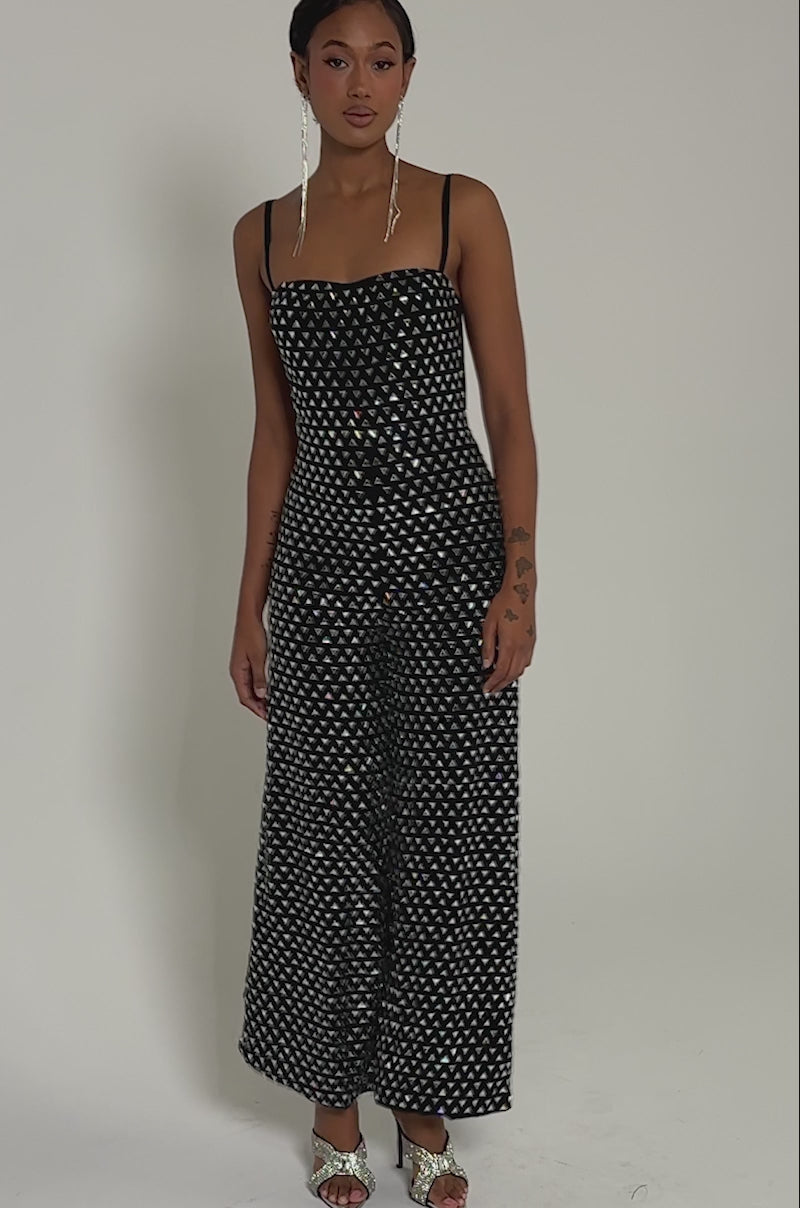 Video of woman modeling the CLARA RHINESTONE WIDE LEG JUMPSUIT, a black party piece with a bold white triangle design. She completes her look with statement earrings and strappy metallic heels against a plain gray background.