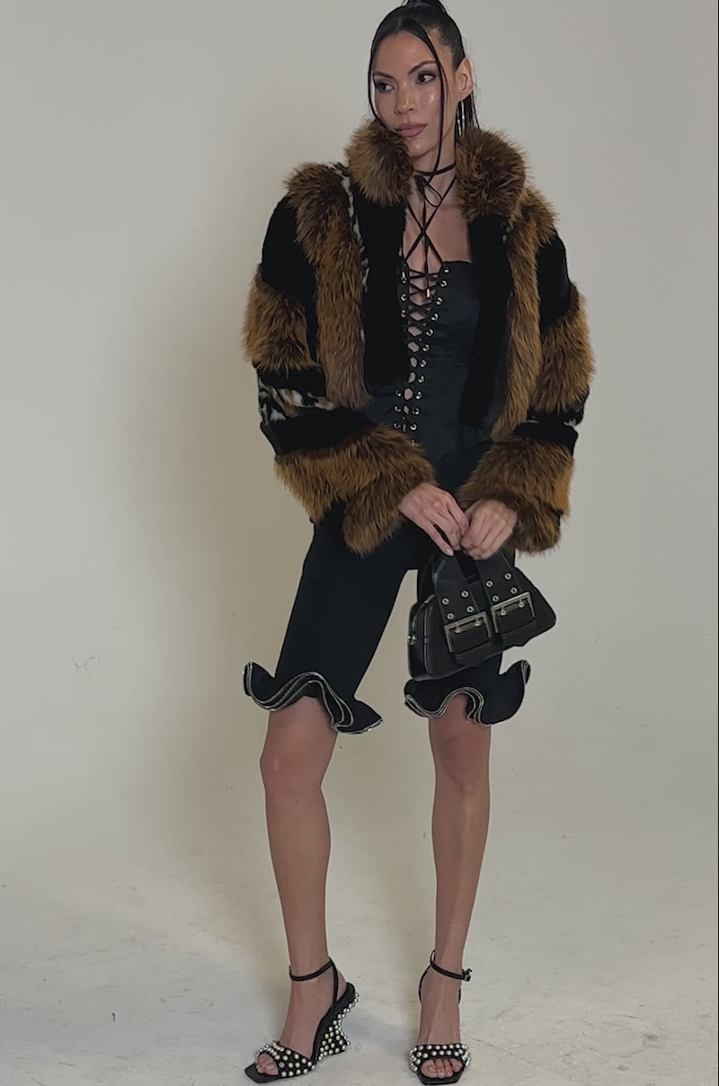 A video of a woman posing confidently in DAINTY DETAILS RUFFLE HEM TAILORED SHORTS, a black lace-up corset top, brown and black faux fur jacket, high-heeled sandals, and a small black handbag.