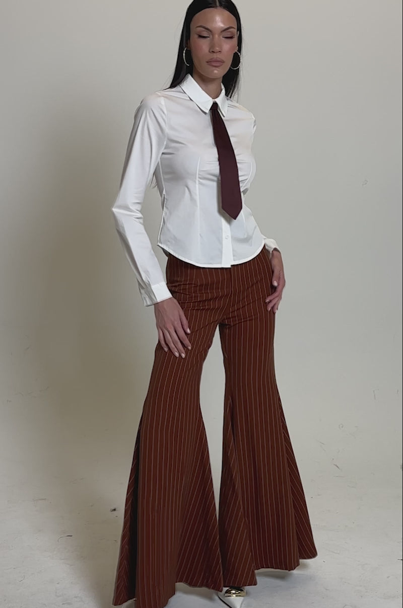 A video of a woman wearing the BUSINESS FIRST BUTTON DOWN blouse with a detachable satin necktie, styled with high-waisted rust pinstripe flared pants and pointed-toe shoes, posing against a plain light background.