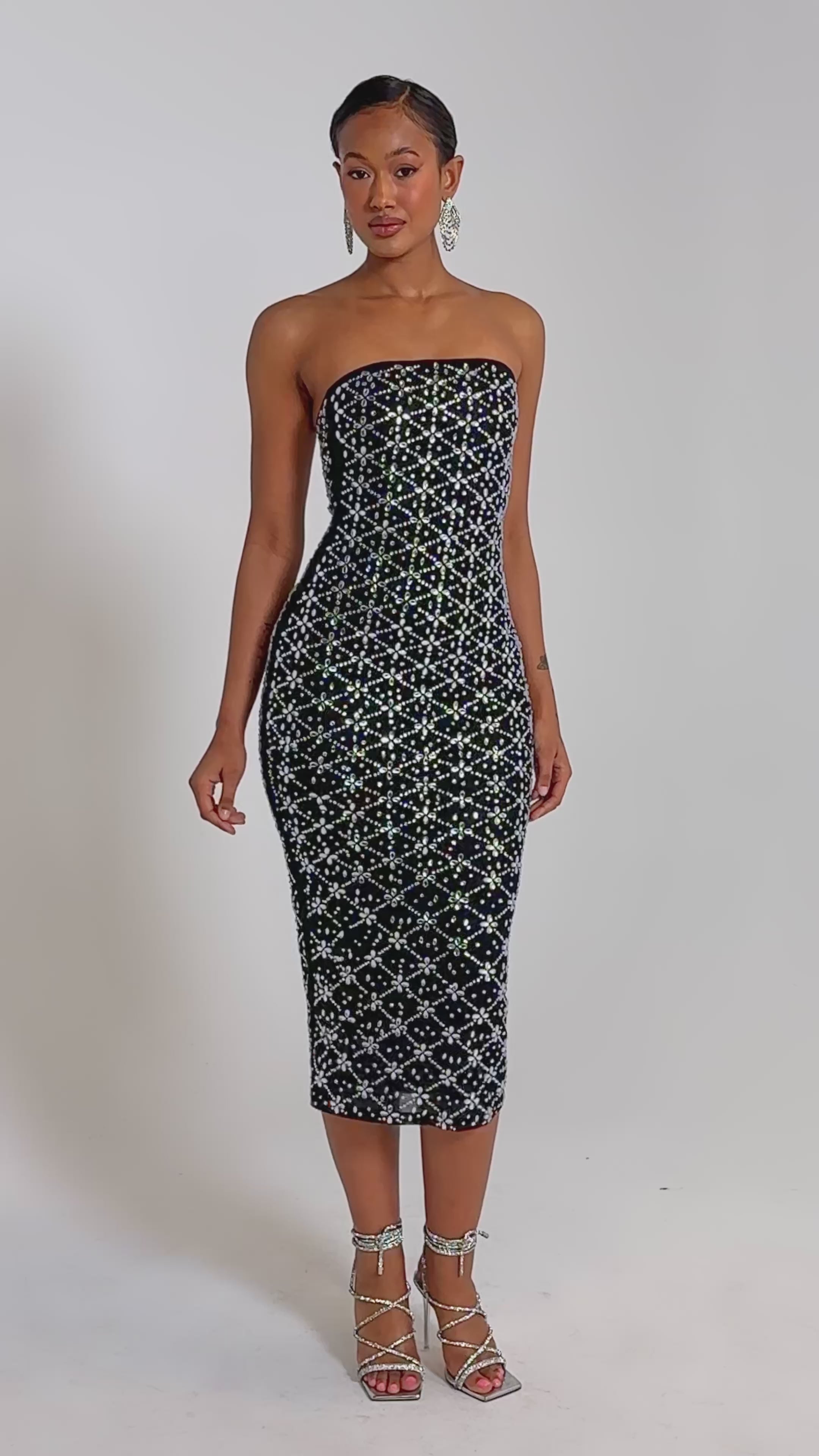 A video of a woman in the MORE THE MERRIER EMBELLISHED MAXI DRESS, a strapless, body-contouring black dress with white and silver sequin details, paired with silver strappy heels.