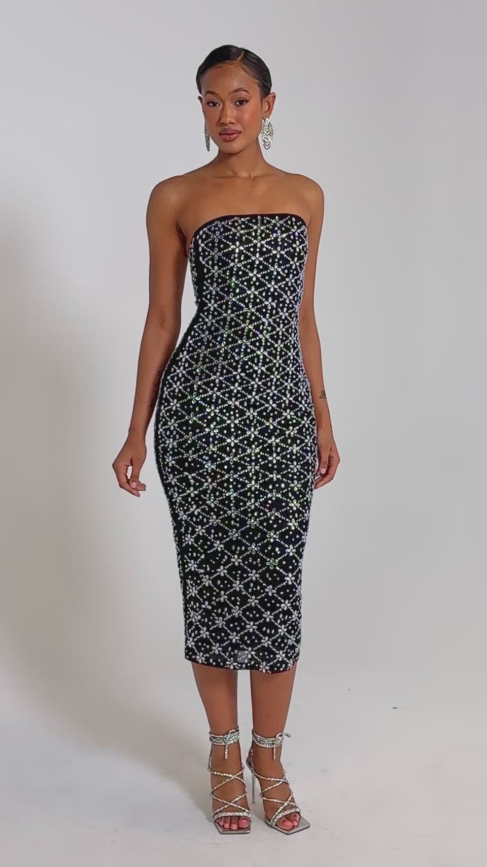 A video of a woman in the MORE THE MERRIER EMBELLISHED MAXI DRESS, a strapless, body-contouring black dress with white and silver sequin details, paired with silver strappy heels.