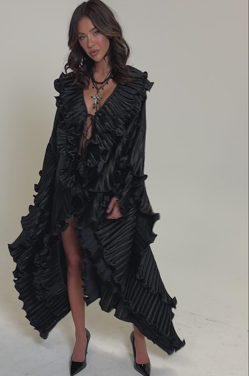 Video of woman modeling the SHINE ON LONG SLEEVE FRONT TIE TOP styled as a chic black dress with oversized sleeves, deep V-neckline, and black heels against a plain light background.