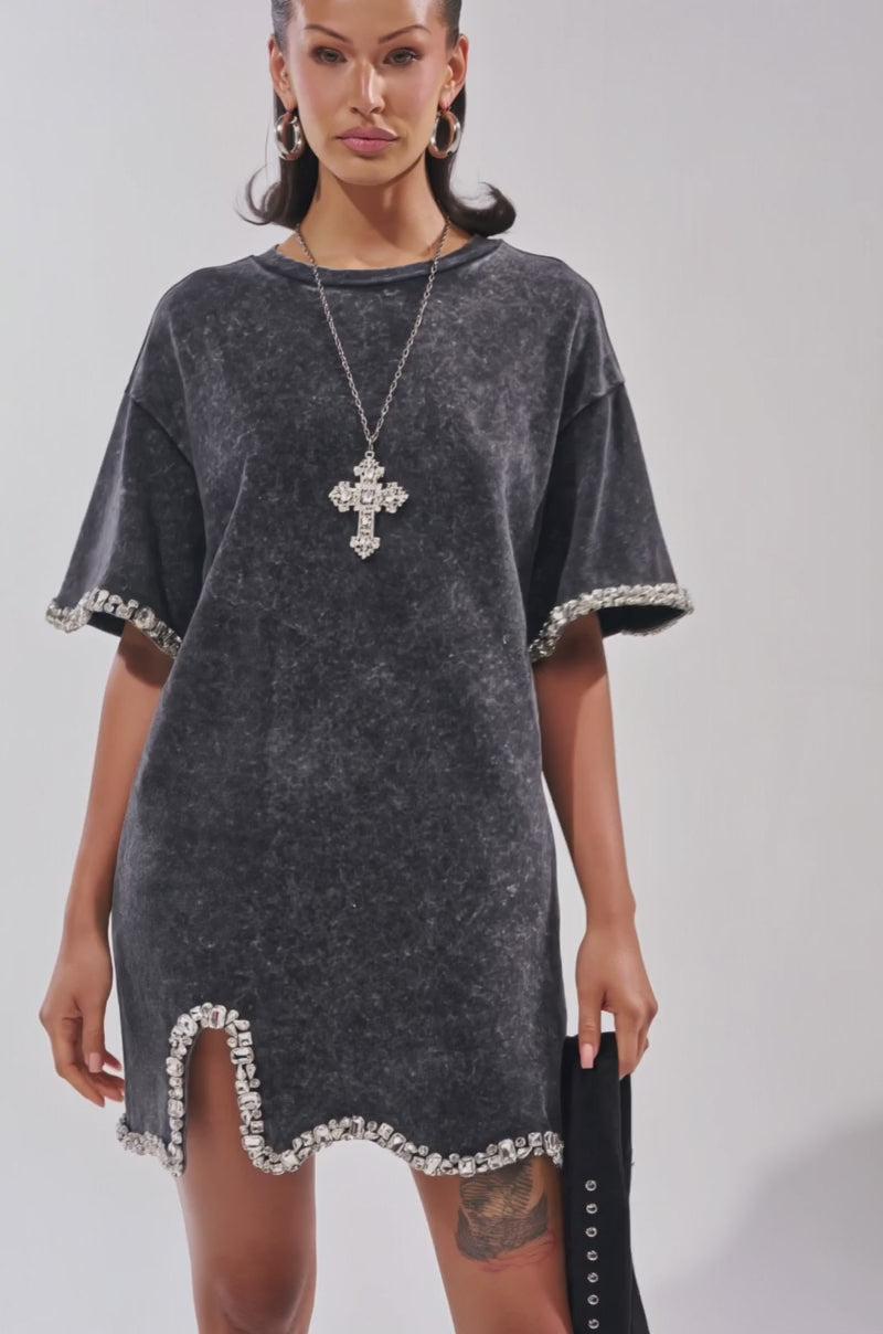 Video of woman modeling the COOL GIRL EMBELLISHED TEE SHIRT DRESS IN BLACK with a cross necklace, hoop earrings, fringe heeled boots, and a black bag. 