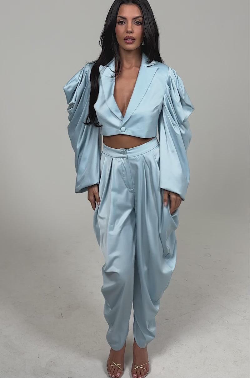 Video of woman modeling the DRIP DRIP TROUSER, a shiny light blue high-rise satin pant paired with a dramatic draped sleeve top, standing in heels against a plain background.
