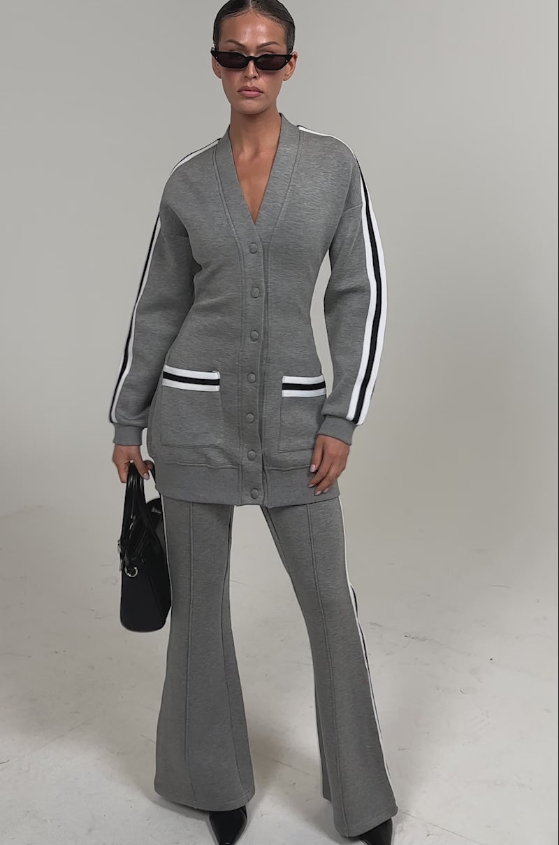 Video of woman modeling the YOU KNOW THE DRILL CARDIGAN with white and black striped pockets, matching flared pants, a black handbag, black sunglasses, and heeled boots for sporty street style.