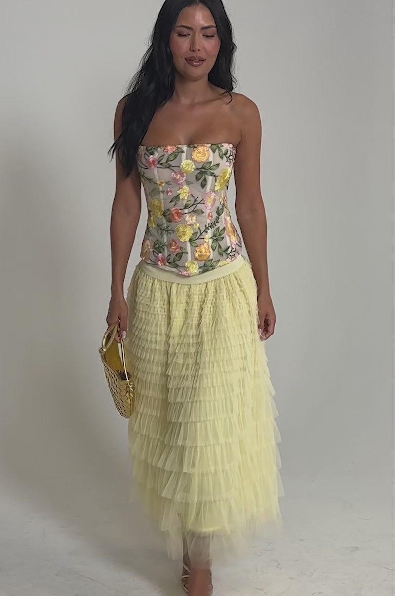 Video of woman modeling the STARS ALIGNED MESH RUFFLE MAXI SKIRT IN BABY YELLOW, featuring tiered ruffles, paired with gold strappy heels and a gold textured handbag. The skirt’s voluminous layers create a standout look.