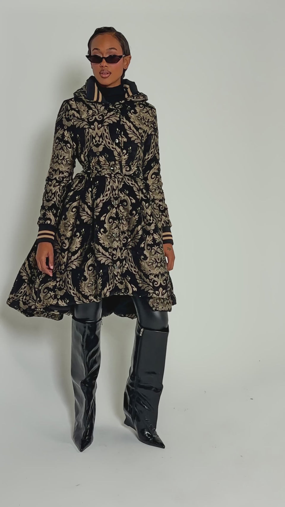 Video of woman modeling the DECKER PUFFER TRENCH IN JACQUARD, featuring an ornate black and gold pattern with a wide collar, striped trim, and belted waist.
