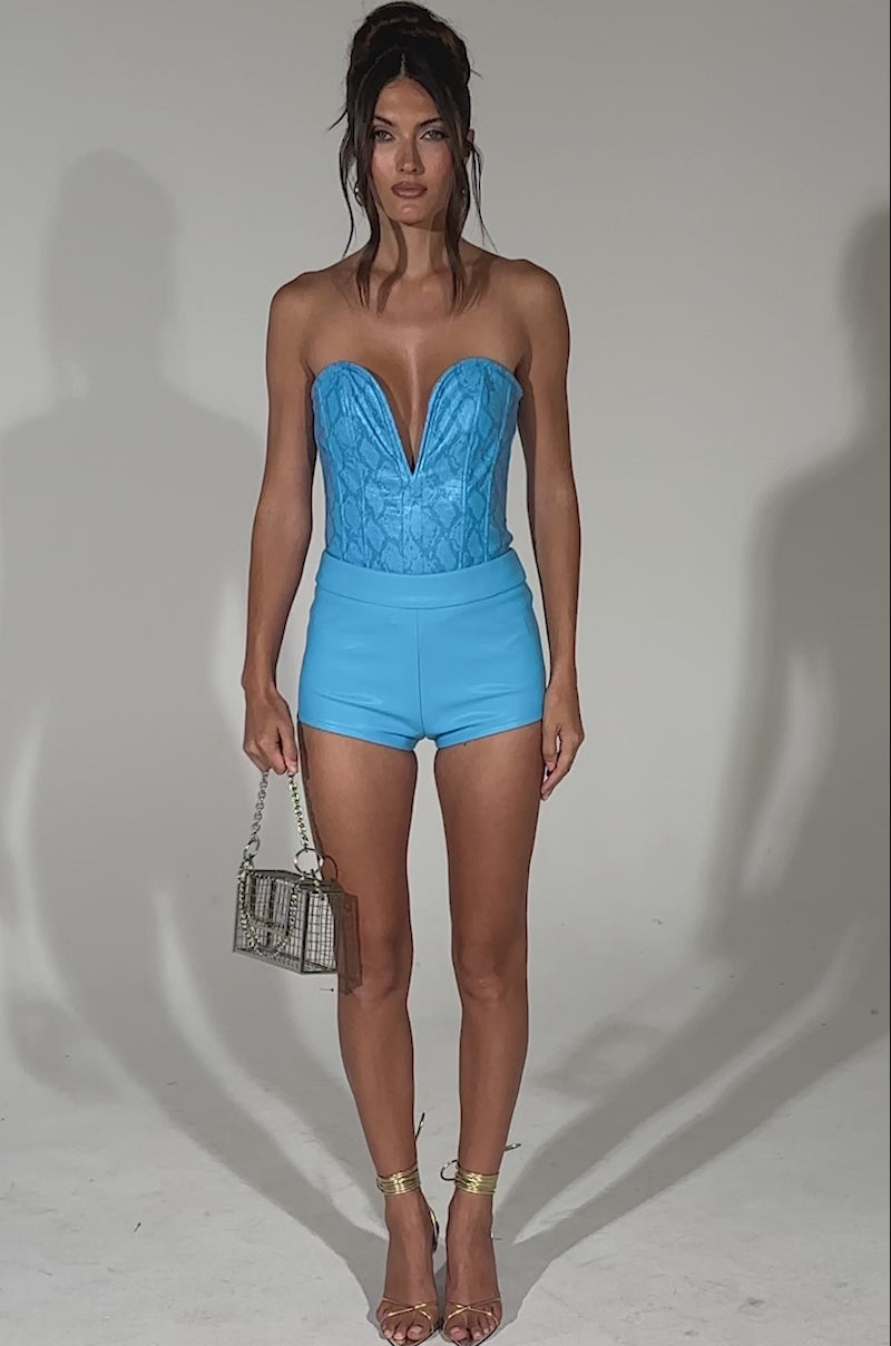 A video of a woman posing against a light background wearing the TOO HOT FAUX LEATHER MINI SHORT IN BLUE, styled with a matching blue strapless top, gold heels, and a metallic purse.