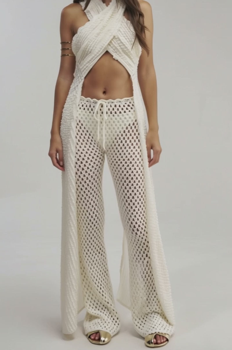 Video of woman modeling the CIAO BELLA CRISS CROSS TOP in cream with matching wide-leg, see-through crochet pants featuring a drawstring waist. She has long hair and gold open-toe shoes.