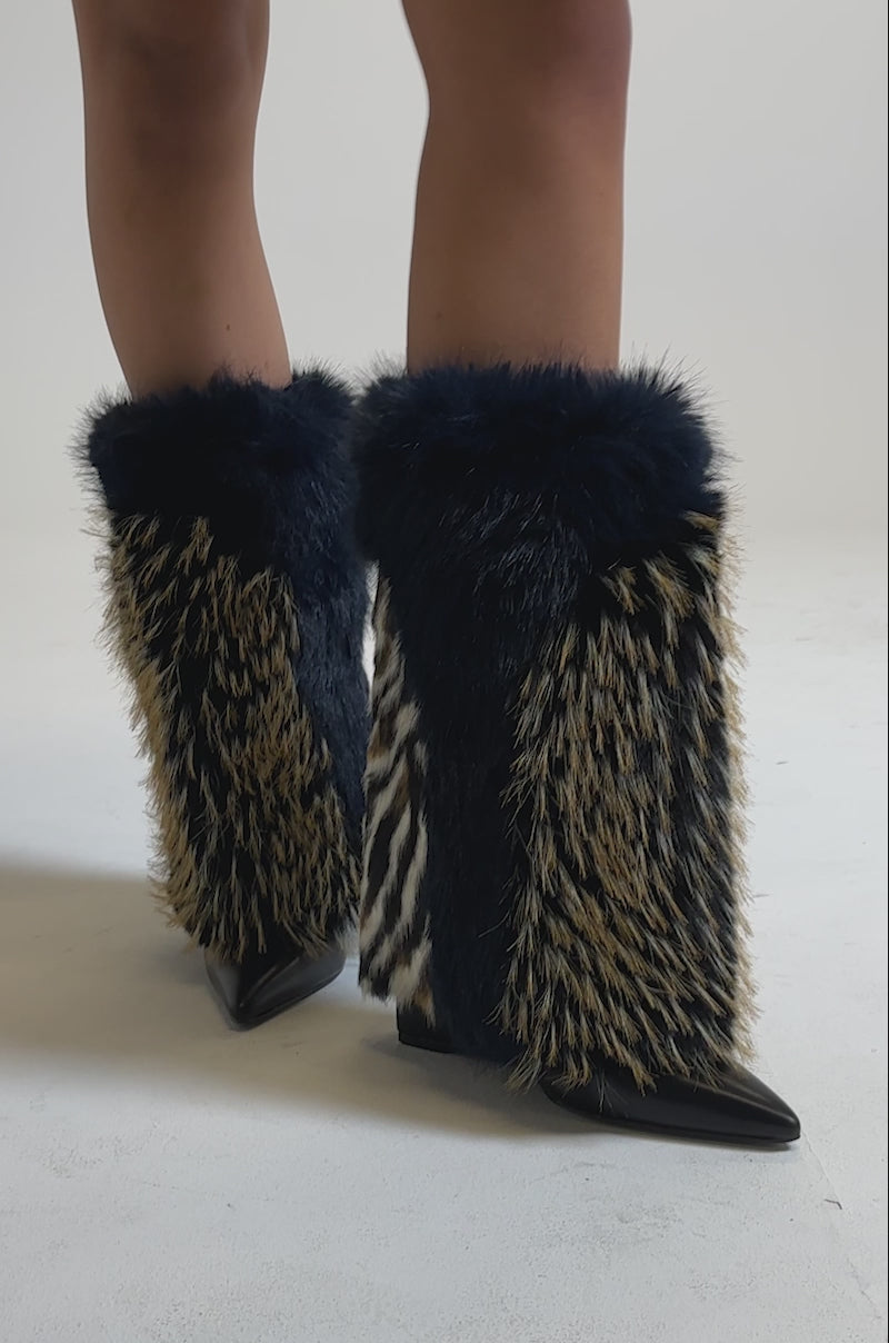 A video of someone wearing the AZALEA WANG BRIASH NAVY MULTI WEDGE BOOTIE, featuring black pointed toes with thick faux fur in navy, tan, and animal prints, paired with a shiny burgundy skirt.
