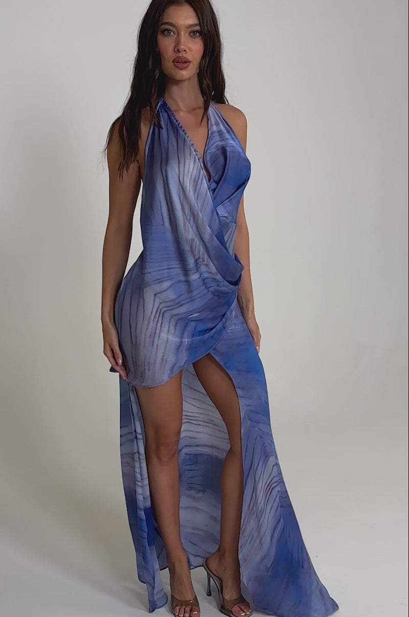 A video of a woman wearing the PLAY NICE MINI DRESS IN PURPLE MULTI, featuring a sleeveless design, purple and white abstract pattern, draped front, high slit, and sheer fabric.