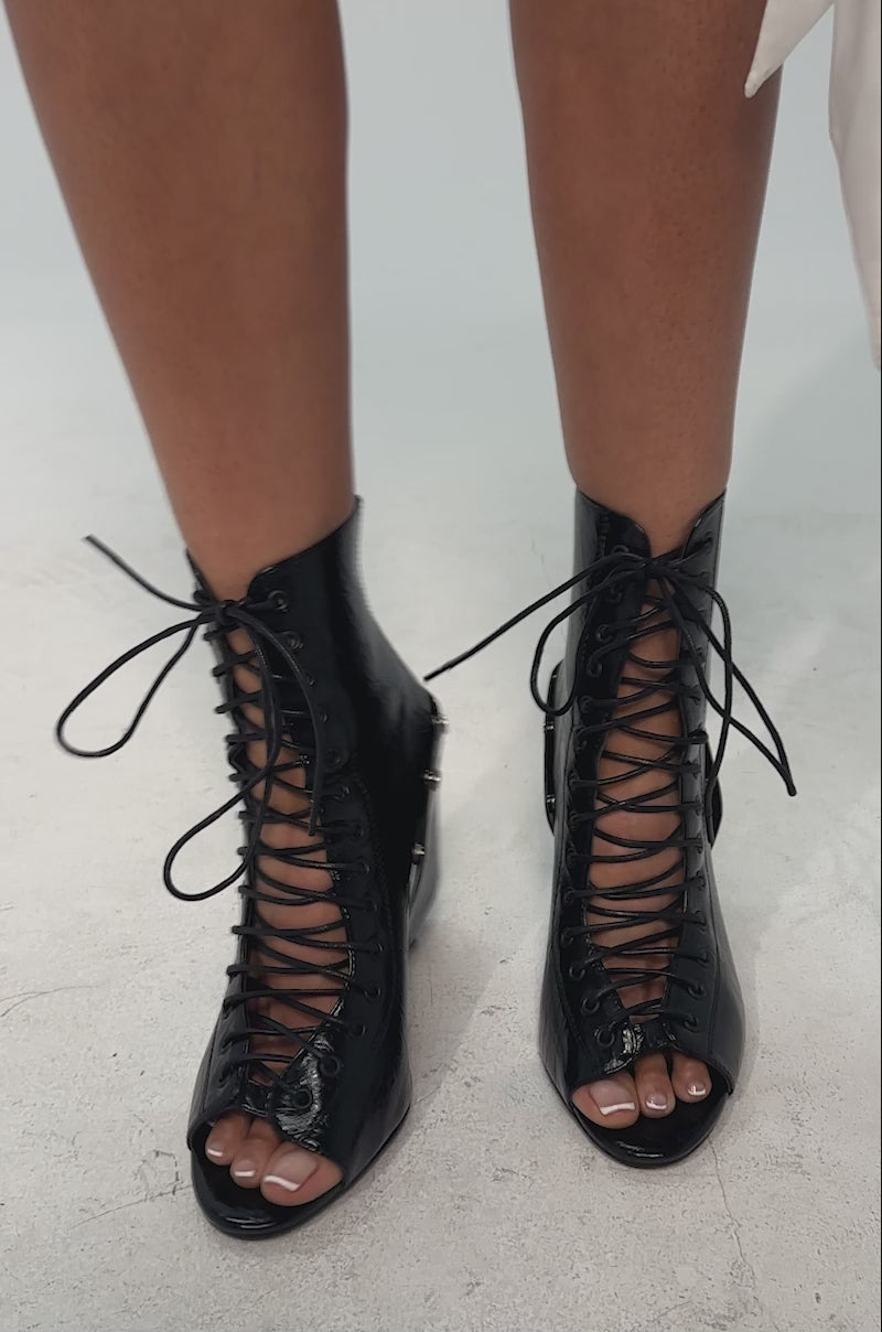 A video of a model wearing the AZALEA WANG ALLURE Black Chunky Lace Upper Open Toe Bootie which features black faux leather, a glossy finish, lace-up ankle, open toe, metallic rivet accents, and a unique chunky block heel.