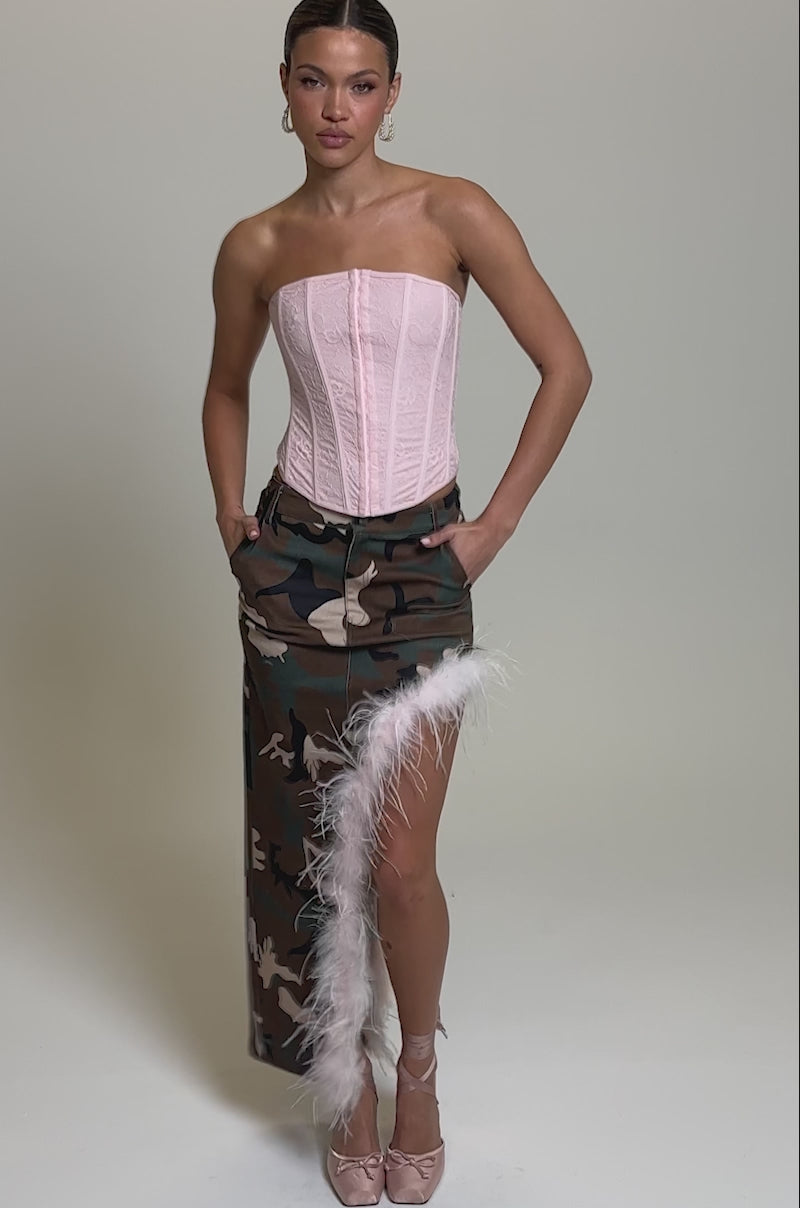 Video of woman modeling the TRYING SOMETHING NEW CAMO SKIRT WITH FEATHERS, pairing the camo midi skirt with feather trim with a strapless pink corset top and pink strappy heels, posing against a plain light background.