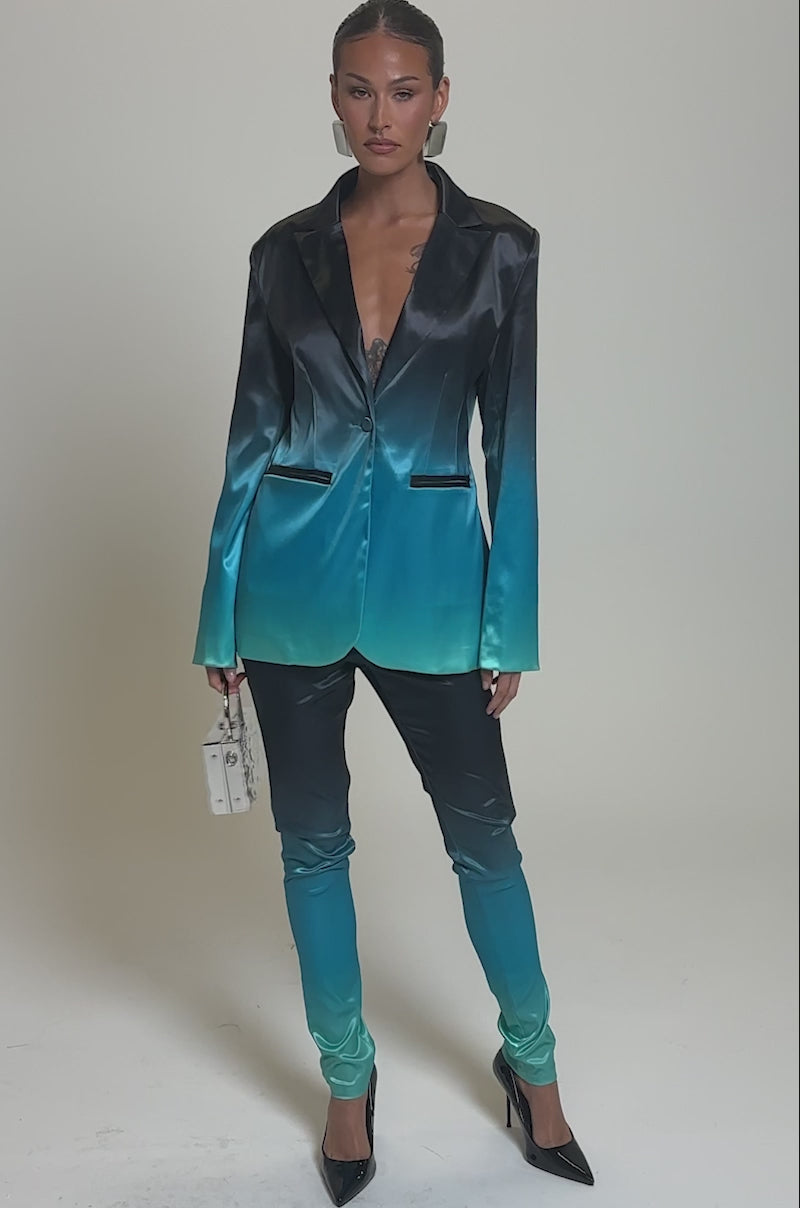 Video of woman modeling the NIGHT AND DAY OMBRE BLAZER and matching pants, which fade from black to teal, she styles the look with a small white handbag and bold silver earrings, posing against a plain background.