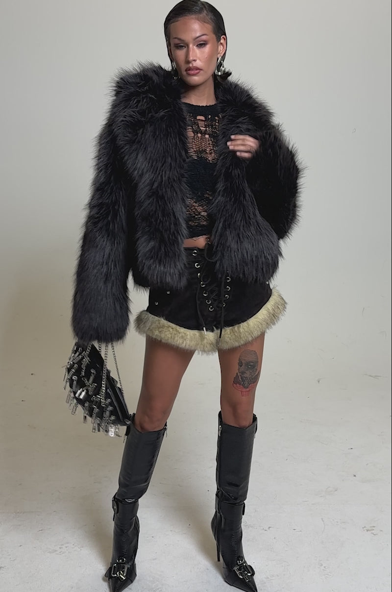A video of a woman in the DUSK TIL DAWN FAUX FUR COAT, paired with a black lace top, fur-trimmed dark shorts, knee-high boots, and a black handbag. She sports slicked-back hair, hoop earrings, and a thigh tattoo.