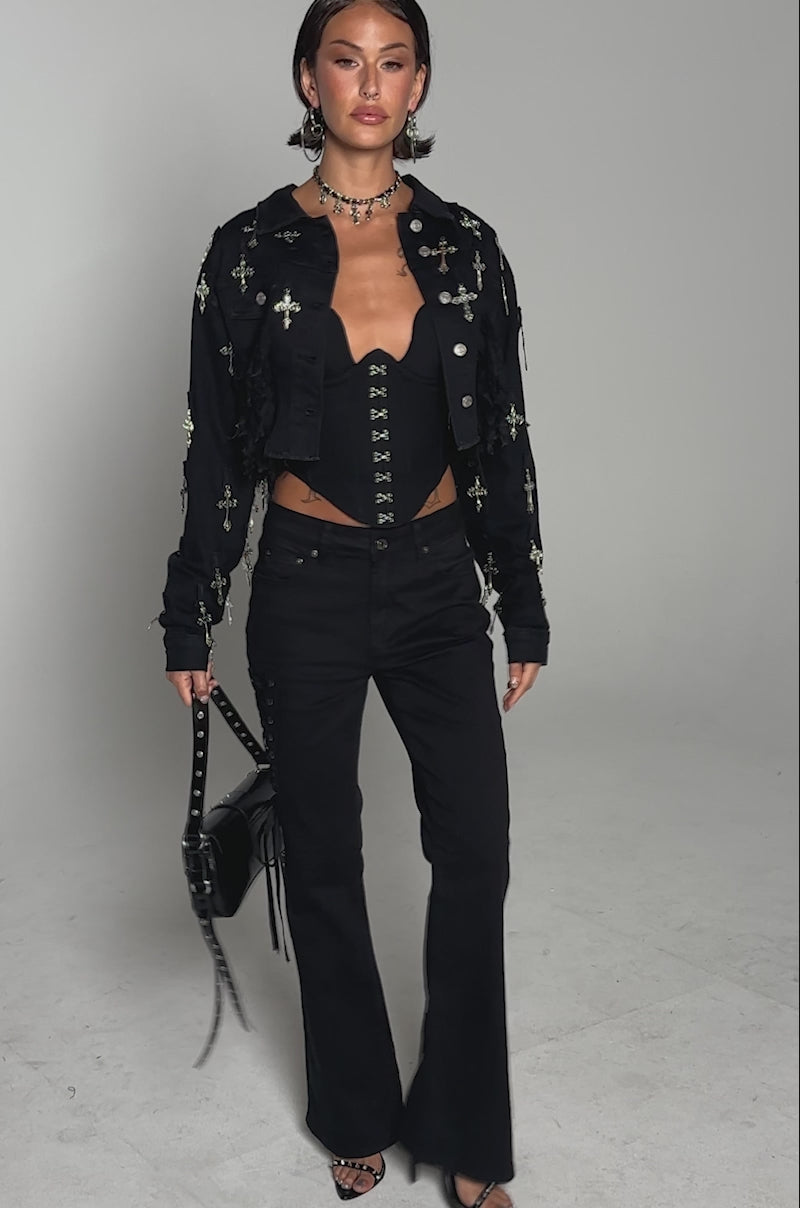 A video of a model wearing the CROSS MY HEART ULTRA CROP DENIM JACKET, a black corset top, black lace-up flared pants, and holding a studded handbag while standing confidently against a plain background.