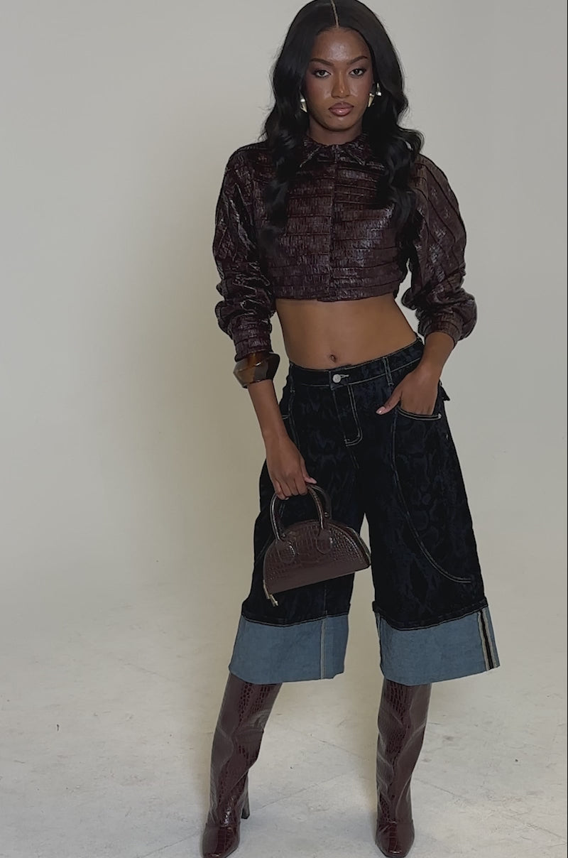 A video of a woman posing in the JUST LIKE THAT BUTTON DOWN cropped shirt, dark wide-leg denim shorts with light cuffs, maroon knee-high boots, and holds a small brown handbag against a plain background.
