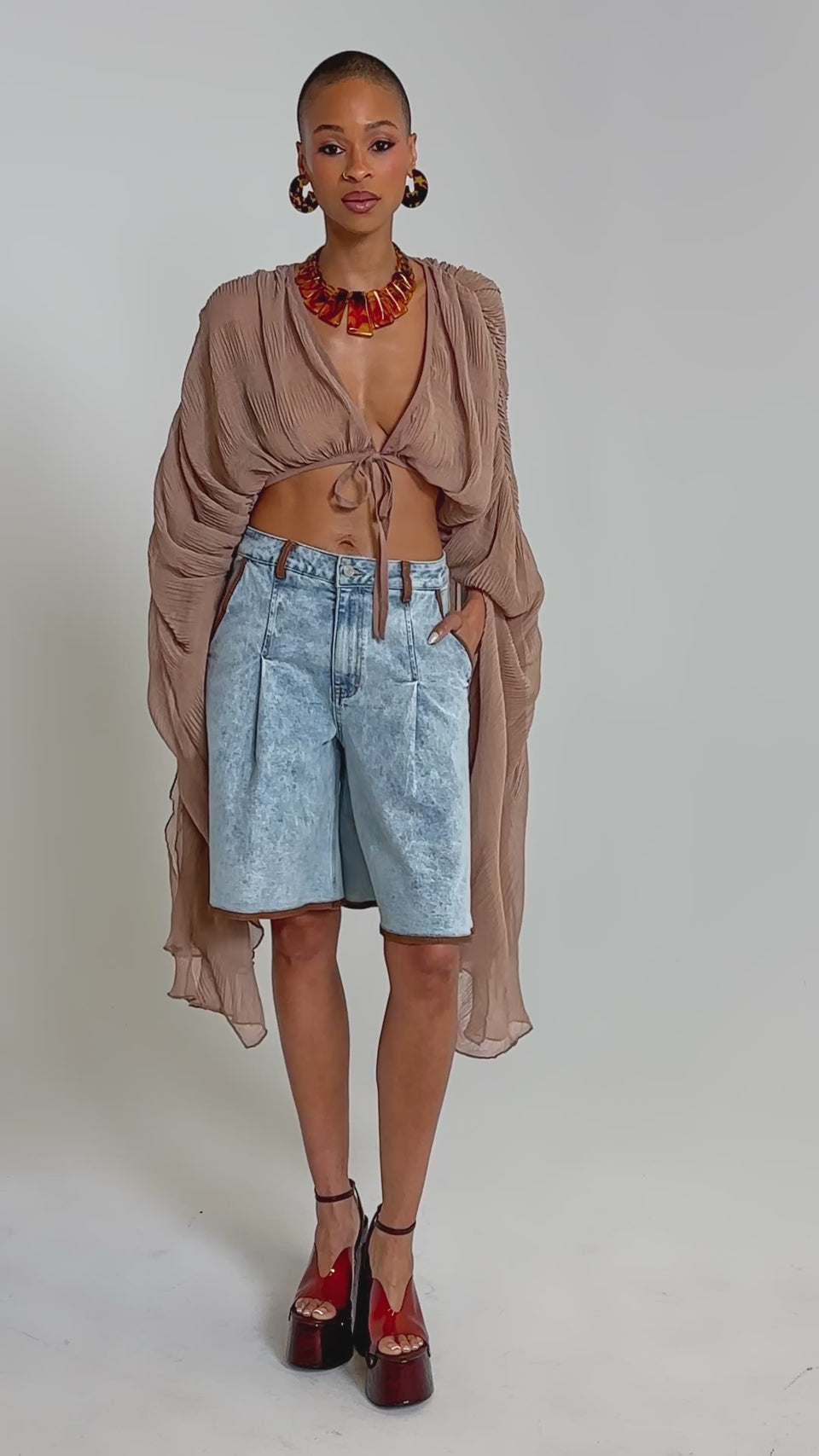 Video of woman modeling the FLY AWAY CHIFFON BLOUSE IN TAUPE, featuring sheer fabric, wide sleeves, and a tie front, styled with high-waisted denim shorts. She accessorizes with bold earrings and a chunky necklace against a plain background.