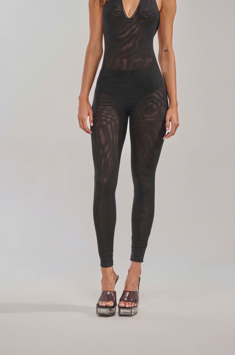 Video of woman modeling the KEEPIN IT SIMPLE MESH SLEEVELESS JUMPSUIT IN BLACK. She completes the look with layered necklaces and platform heels against a plain, light-colored background.