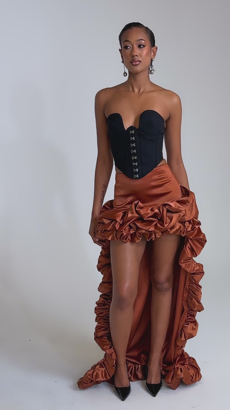 A video of a woman in a strapless black corset top and the LOOK AT ME GO SATIN HIGH LOW SKIRT with dramatic copper ruffles, styled with black heels and statement earrings against a light gray background.